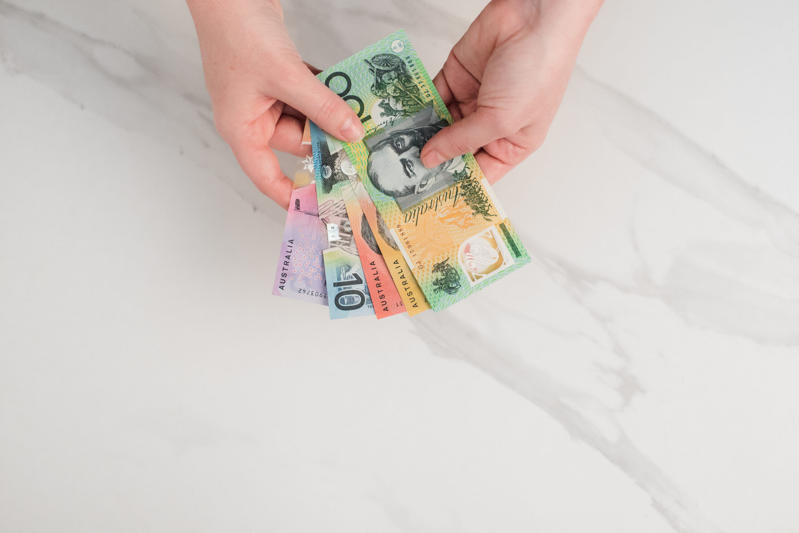 A close-up of a person’s hands holding several Australian banknotes of different denominations, including $5, $10, $20, and $50 notes. The notes are spread out slightly, showing their distinctive colours and designs. The background is a light marble surface, giving the image a clean and professional look that evokes themes of finance, budgeting, or investment.