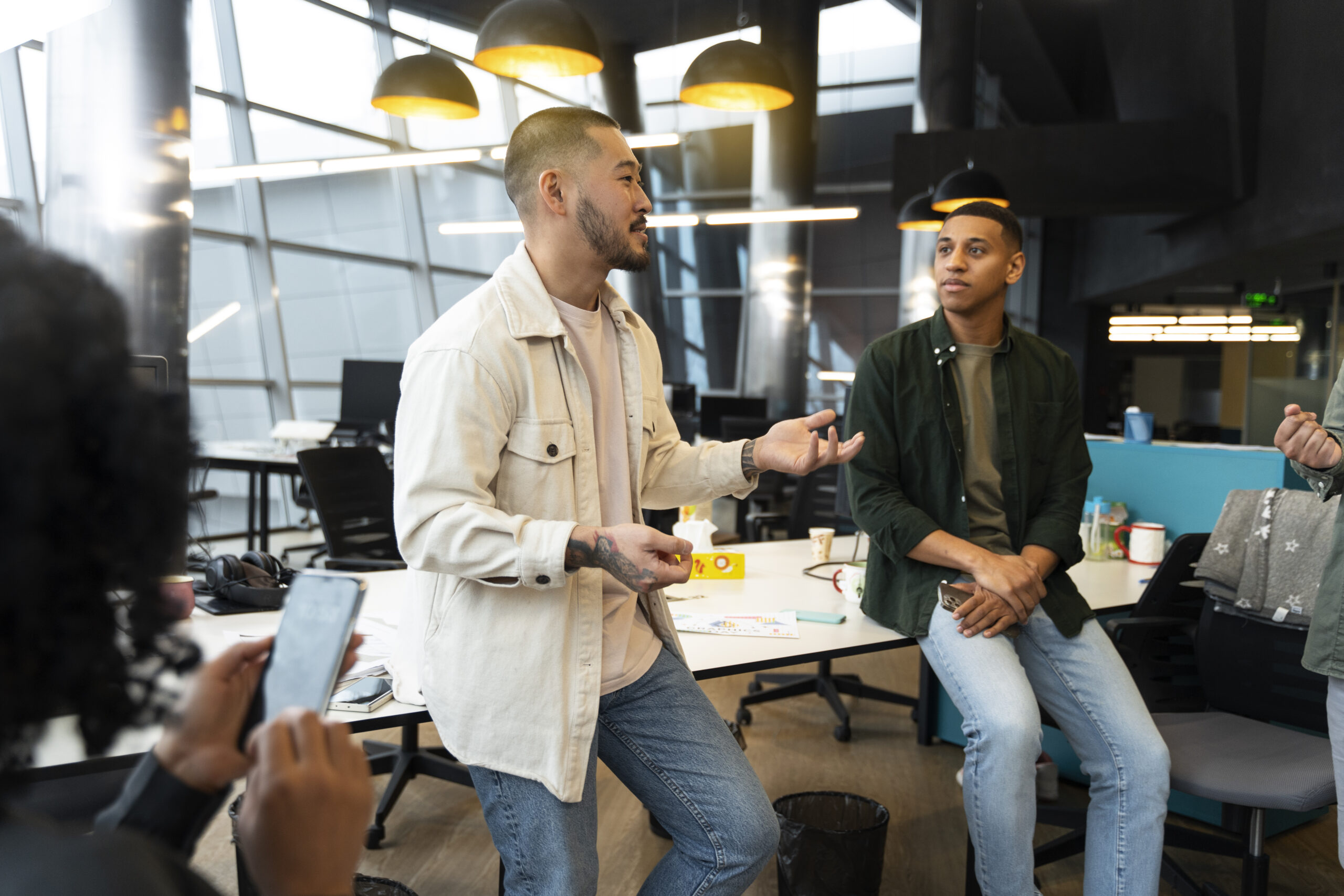 A group of young professionals collaborate in a modern open-plan office with large windows and bright overhead lights. One person stands mid-conversation, gesturing with his hands as he speaks to a colleague sitting casually on a desk. Others listen and engage, with one person holding a smartphone in the foreground. The setting feels informal and creative, suggesting a team meeting or brainstorming session in a contemporary workspace. Australia's research and development landscape is about to change in a big way. The Australian Government has kicked off an independent strategic examination of research and development to overhaul how the country supports innovation. Final recommendations are due by 31 December 2025. For founders, CFOs and emerging sectors like AI and biotech, what happens next could be the difference between extending your runway and running out of cash.