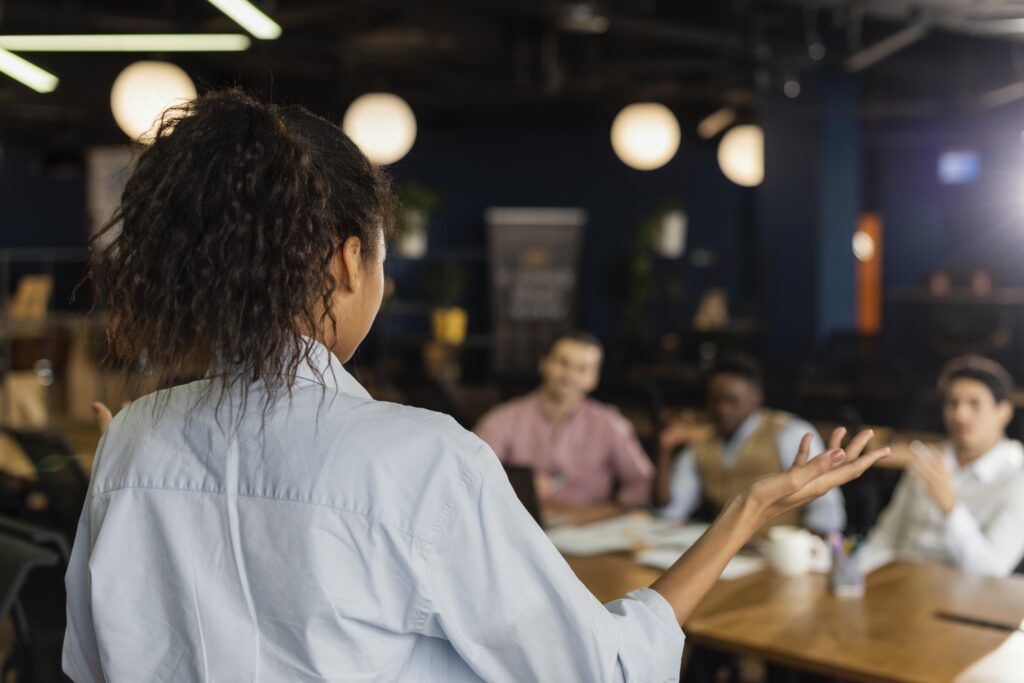 A woman speaks to a small group seated at a table during an informal presentation.
