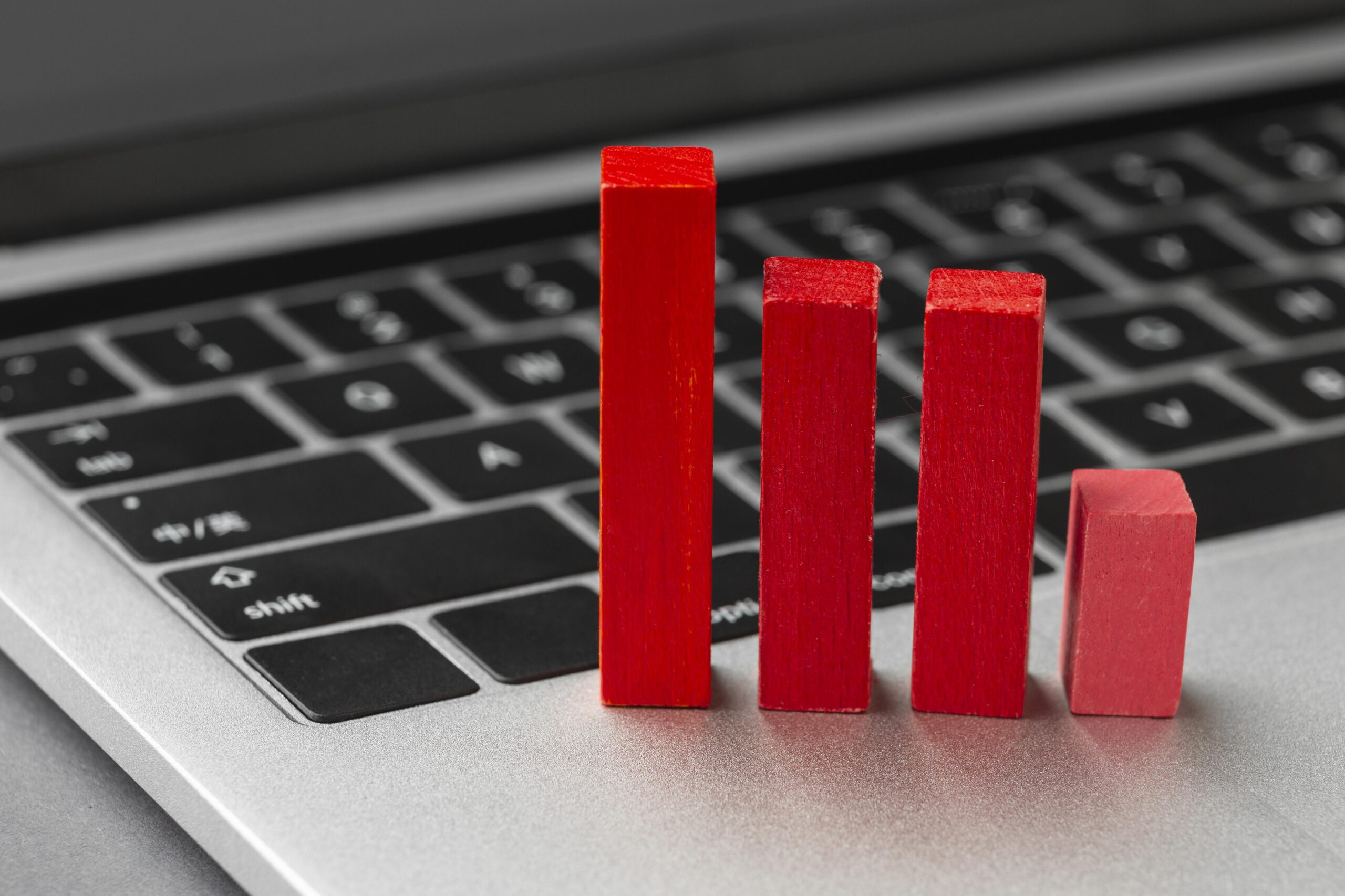 Close-up of a silver laptop with black keys, where four red wooden blocks stand in a row on the keyboard like a bar chart, descending in height from left to right to symbolise a drop in metrics, revenue, or R&D performance.