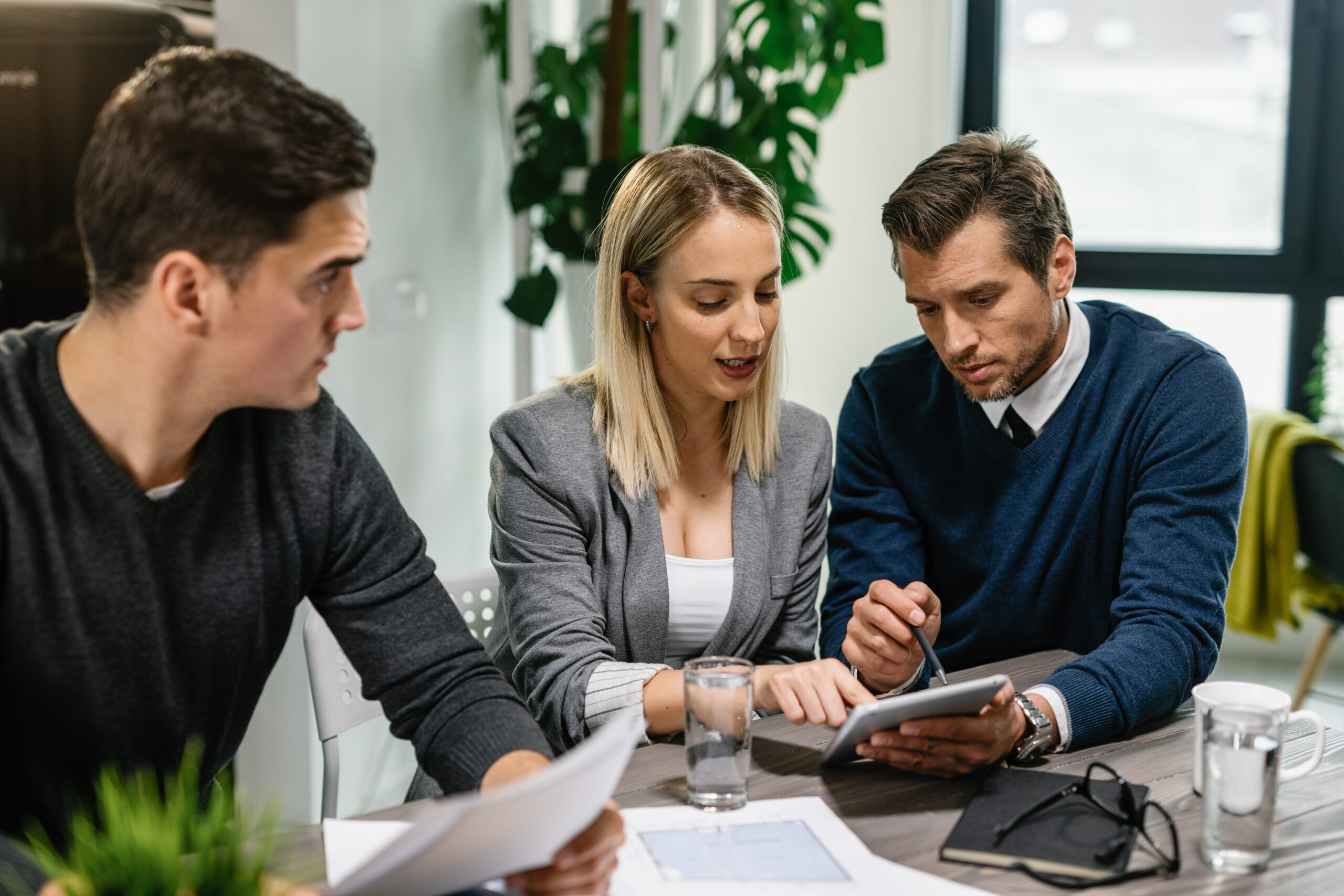 Three business professionals sit together at a meeting table in a modern office. A woman in a light grey blazer is seated between two men. She is looking at a digital tablet while one man, dressed in a navy sweater over a collared shirt, points at the screen to explain something. The other man, wearing a dark jacket, holds printed documents and listens attentively. Papers, glasses of water, and a notebook are spread across the table, with large windows and indoor plants visible in the background. The group appears to be discussing financial or business planning.