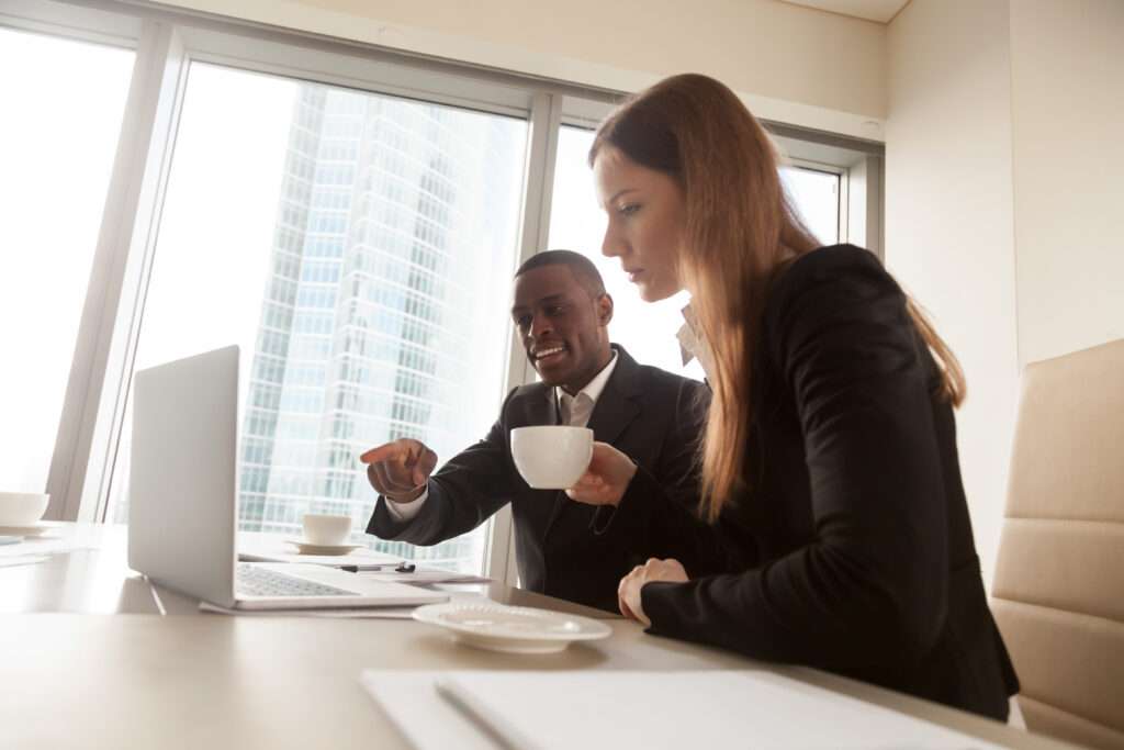 Two professionals, a man with a coffee cup and a woman, looking at and pointing to a laptop screen in a high-rise office.