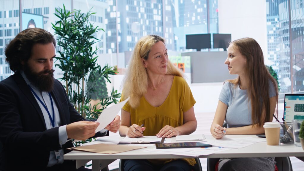 Three colleagues collaborating at a desk. A man with a beard reviews papers while two women discuss the contents.