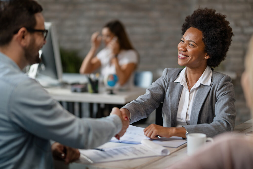 A smiling businesswoman in a grey blazer shaking hands with a man across a desk in a modern, brightly lit office setting.