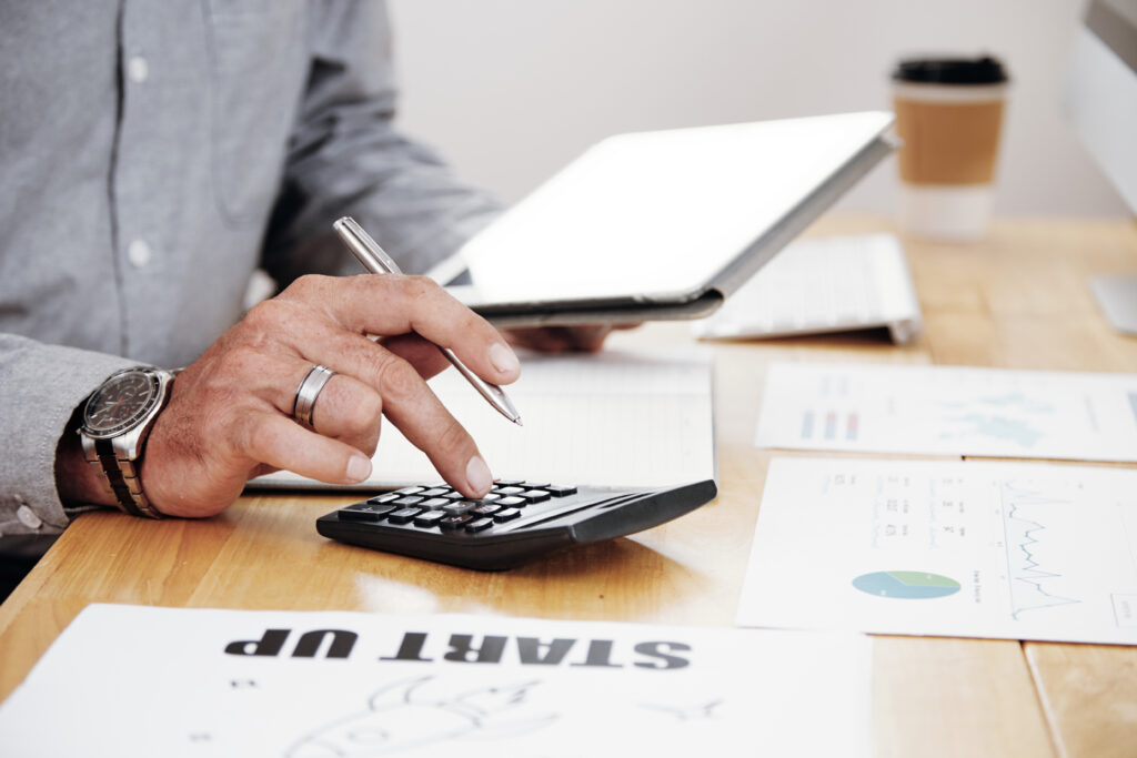 Hands using a calculator next to a tablet and startup business paperwork to calculate an expected R&D tax refund.