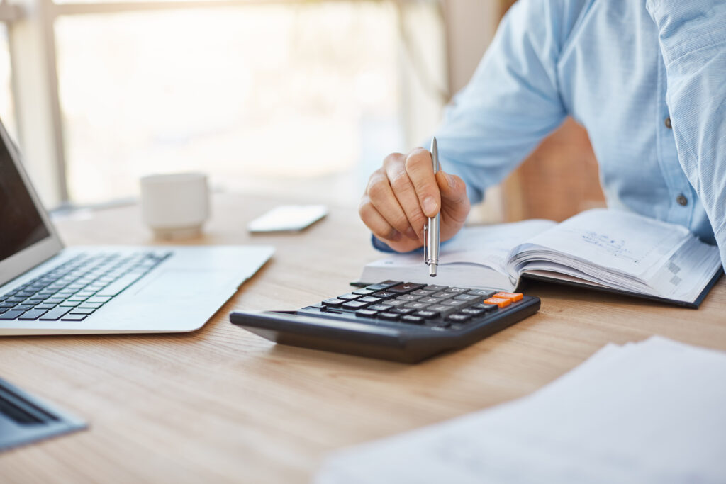 Close-up of hands using a calculator, pen, and notebook next to a laptop to assess tax financials.