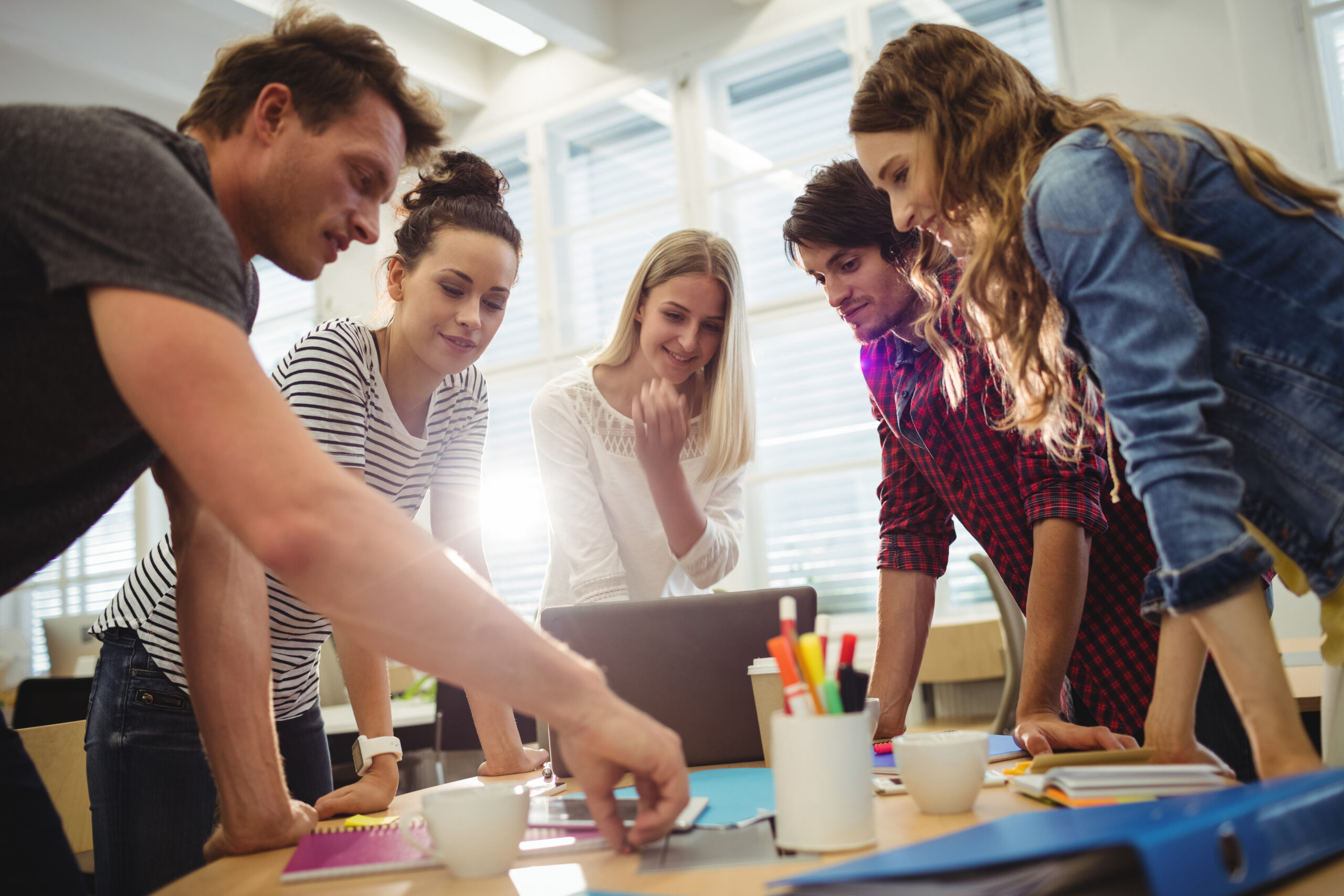 A group of startup founders smiling and collaborating around a laptop and notebooks on a desk in a sunny office.