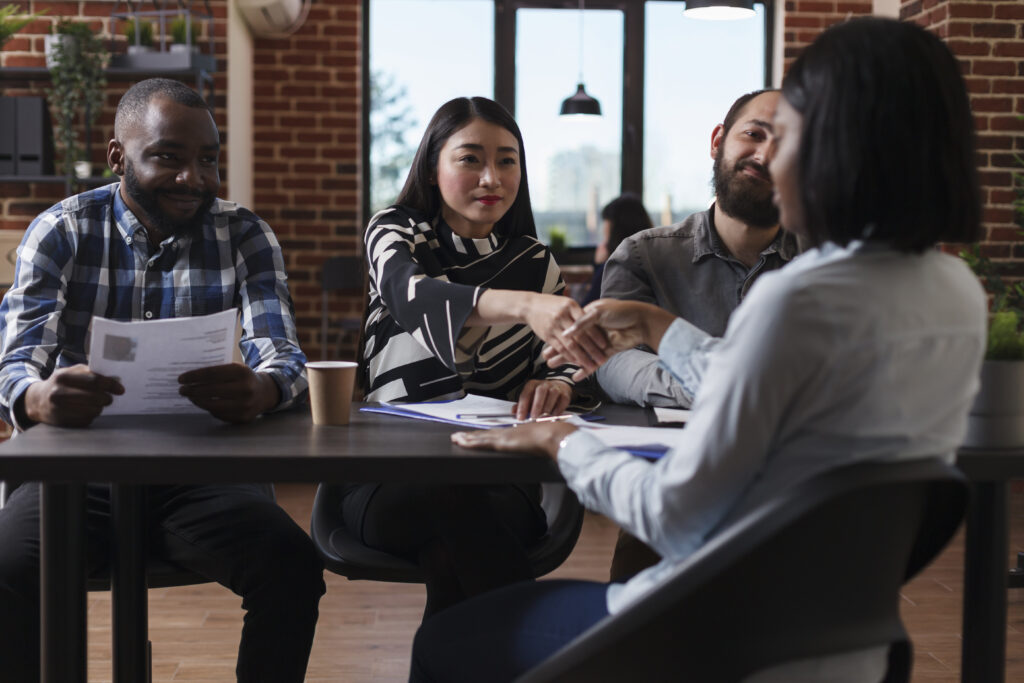Diverse business team smiling as two women shake hands across a desk, finalizing an R&D financing agreement.