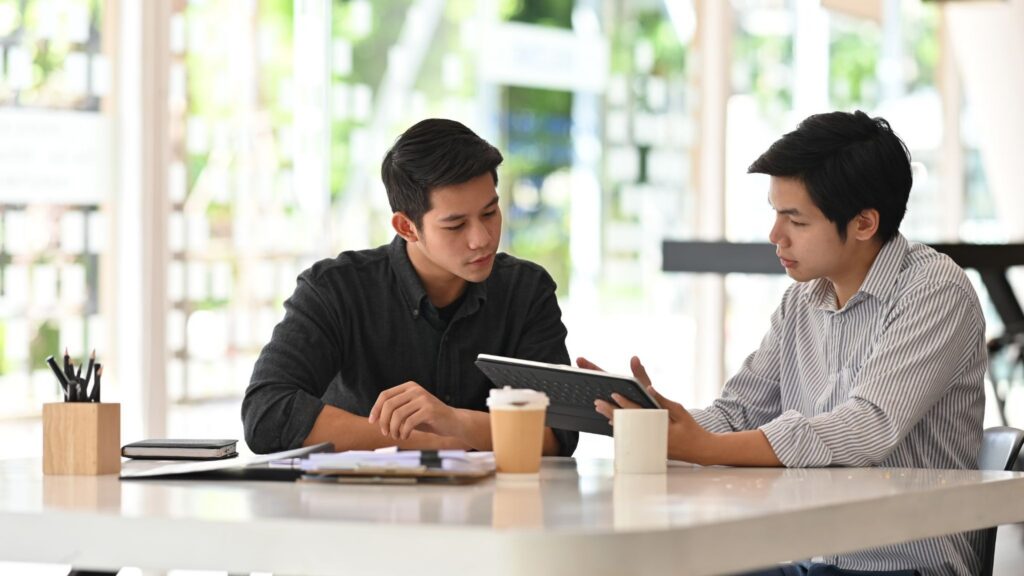 Two men at a desk with coffee discuss R&D finance options and cash flow solutions using a tablet.