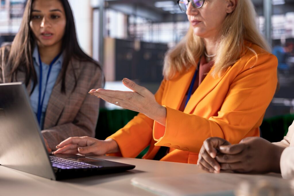 A businesswoman in an orange jacket discusses refundable R&D tax offset calculations on a laptop with a colleague.